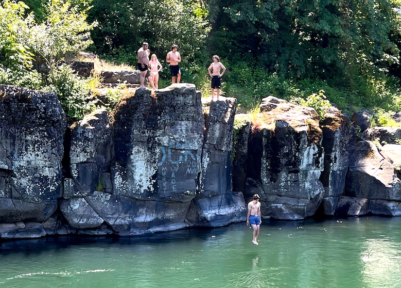 People gather at a rocky outcrop near a body of water and watch as a male jumps into the water.