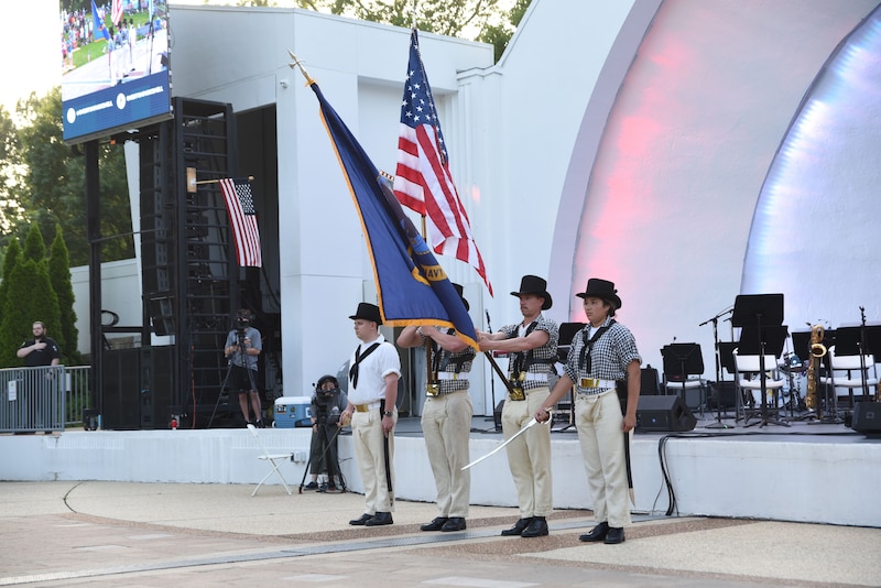 Four sailors in historic garb hold flags and ceremonial swords on an outside stage.
