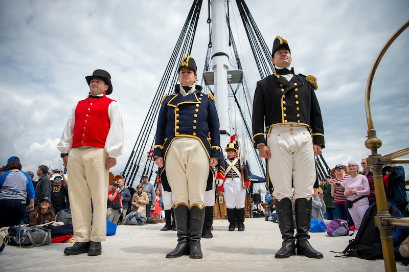 Three sailors in historic dress stand in front of a crowd on an old ship’s deck.