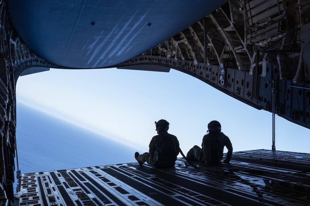 Two seated airmen look out at the blue sky and ocean from the open rear ramp of an airplane.