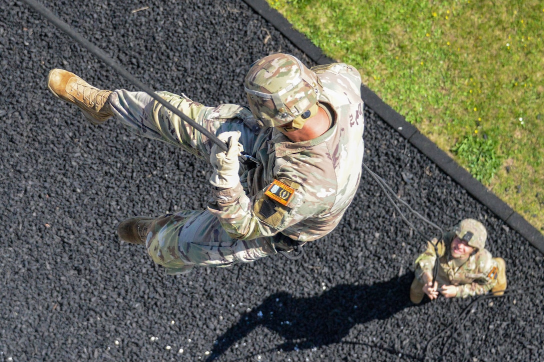 A soldier descends a rappelling rope as another soldier holds the rope and watches on the ground.
