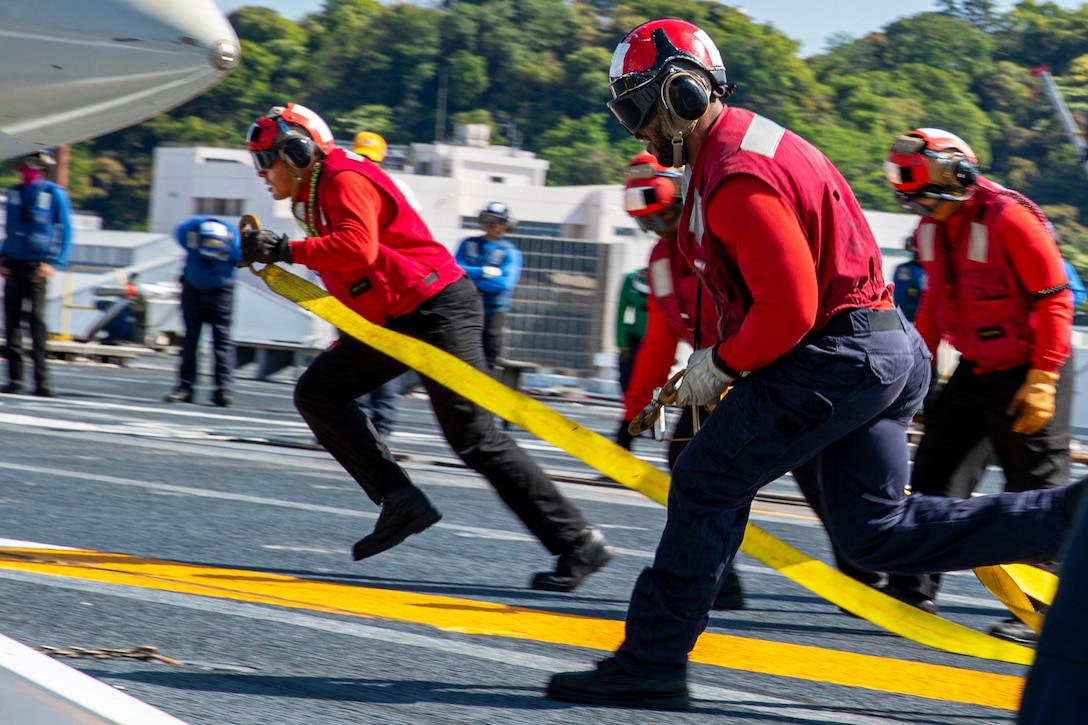 Sailors wearing helmets, goggles and ear protection and carrying a large yellow strap run toward a jet.