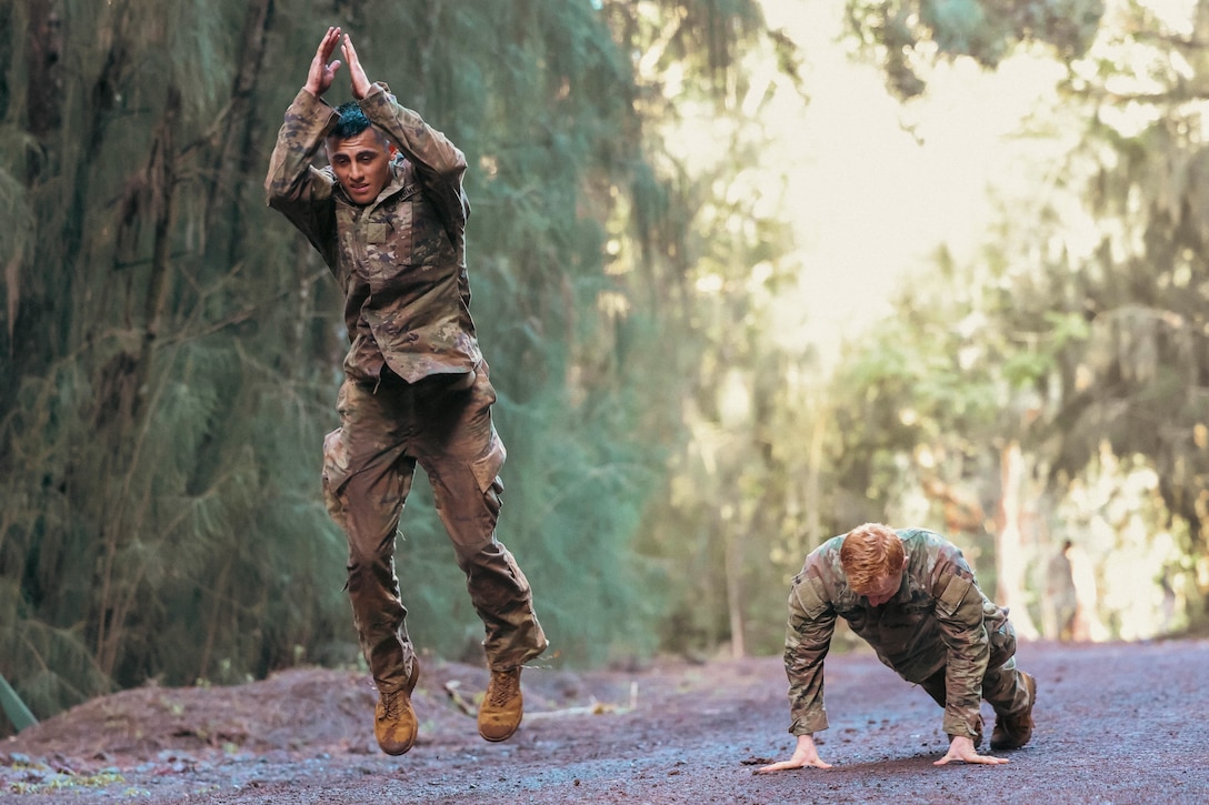 Two soldiers, one jumping and the other in a pushup position, are on a muddy path with trees in the background.