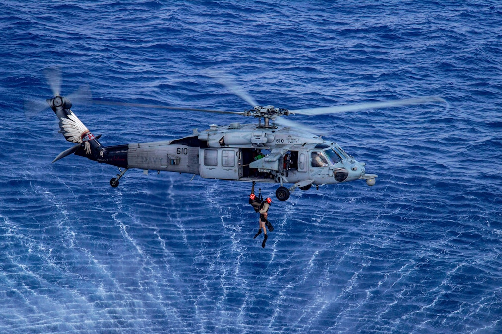 Two sailors attached to a rescue line are lifted into a helicopter from blue water beneath them.