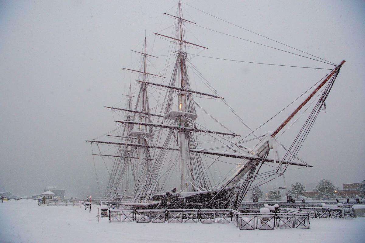 An old ship moored in a harbor is shown during a snowstorm.