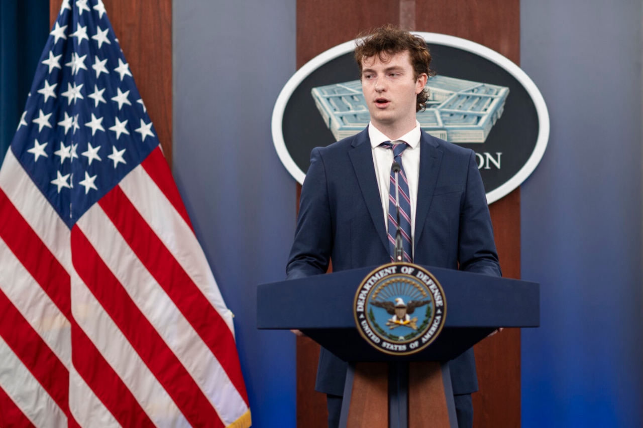 A man stands at a lectern and speaks. To his right is the U.S. flag.