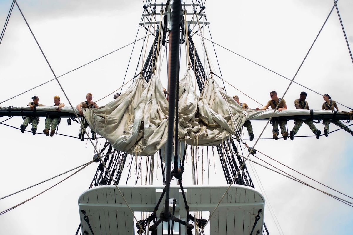 Sailors balance on ropes while holding onto the mast on an old ship.