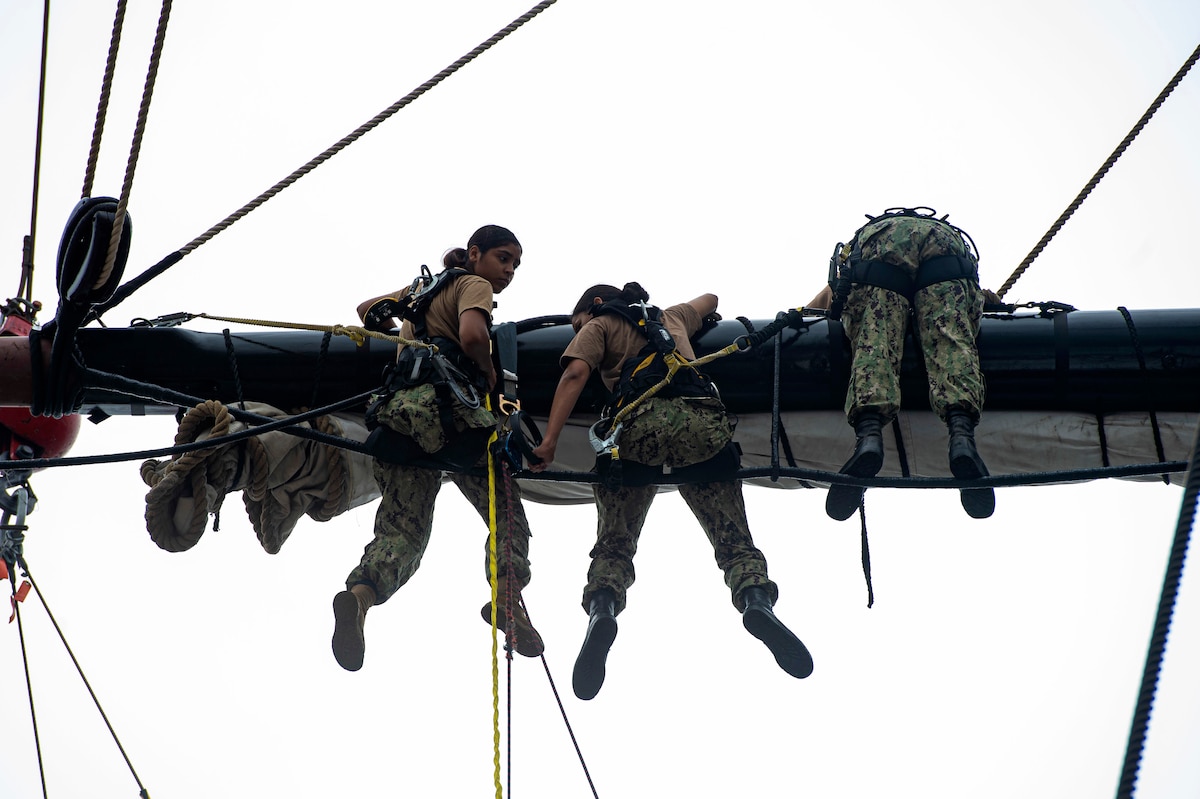 Sailors in harnesses lean over the mast of an old ship.