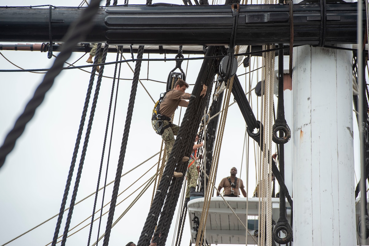 A sailor climbs up the rigging of an old ship while another sailor watches.