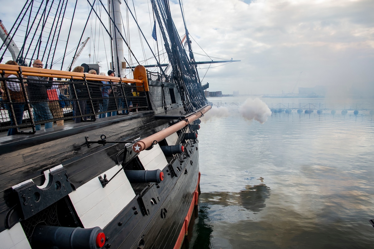 The side of an old ship is shown as smoke comes out of a canon over the harbor.