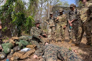 A sailor kneels next to a person lying on the ground as four others stand and watch.