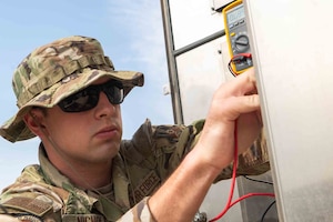 Airman Nichols is to the left of frame with sunglasses on as he's reaching toward the right of frame as he checks the voltage of a Giant Voice System