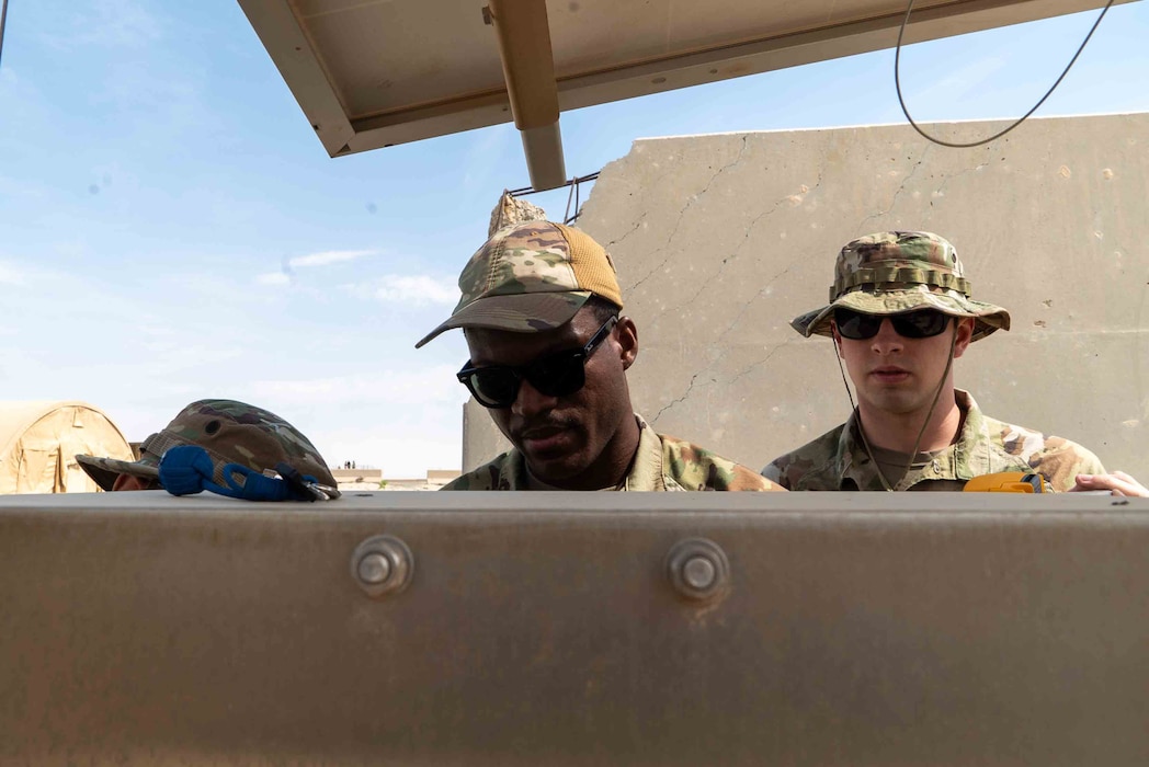 Senior Airman Dadja Dadja stands to the left of Airman Nichols with the panel of a Giant Voice System in the foreground as their heads poke out from the top view