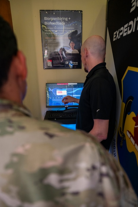 Over the shoulder of an Airman in the foreground, Wyatt Glick with his back turned to camera is showing a demonstration of the MindGym on computer