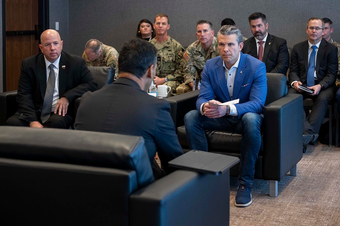 Defense Secretary Pete Hegseth sits in a chair with U.S. personnel behind him and talks with a person seated opposite him.