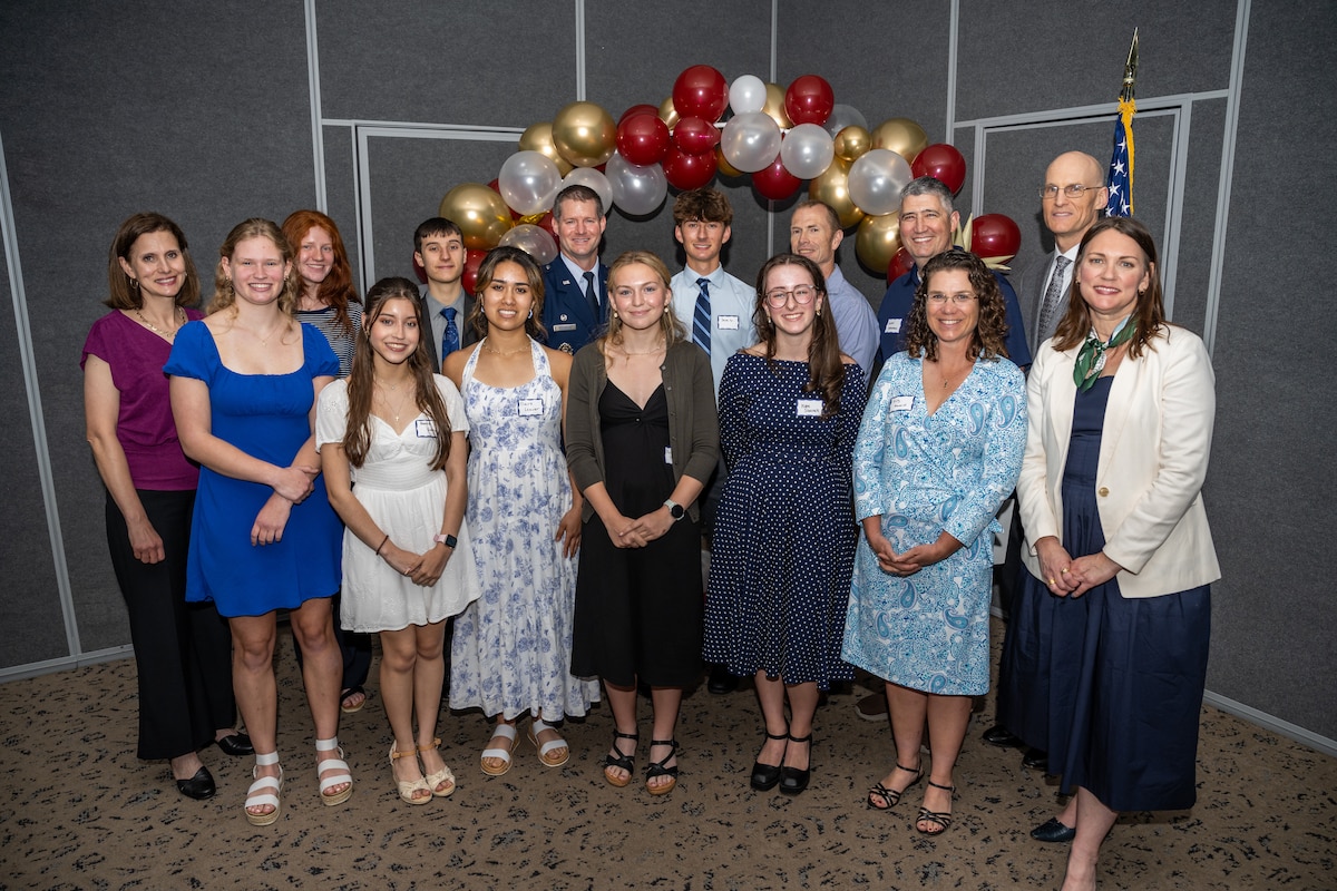 A group photo of 15 people, varying ages, in front of a balloon arch and U.S. flag