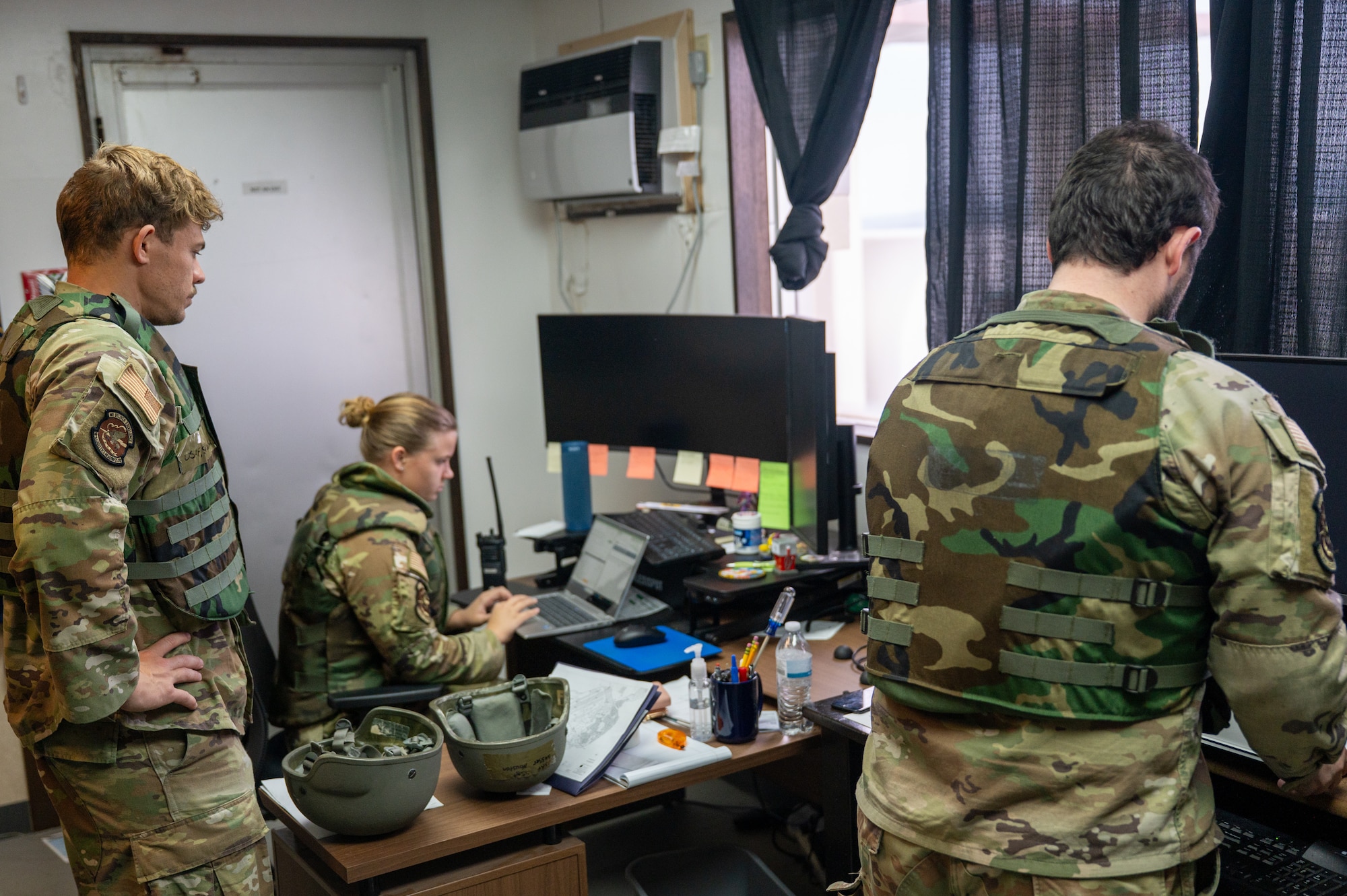 Air Force Airmen sit and stand around a desk while wearing flack vest protective gear