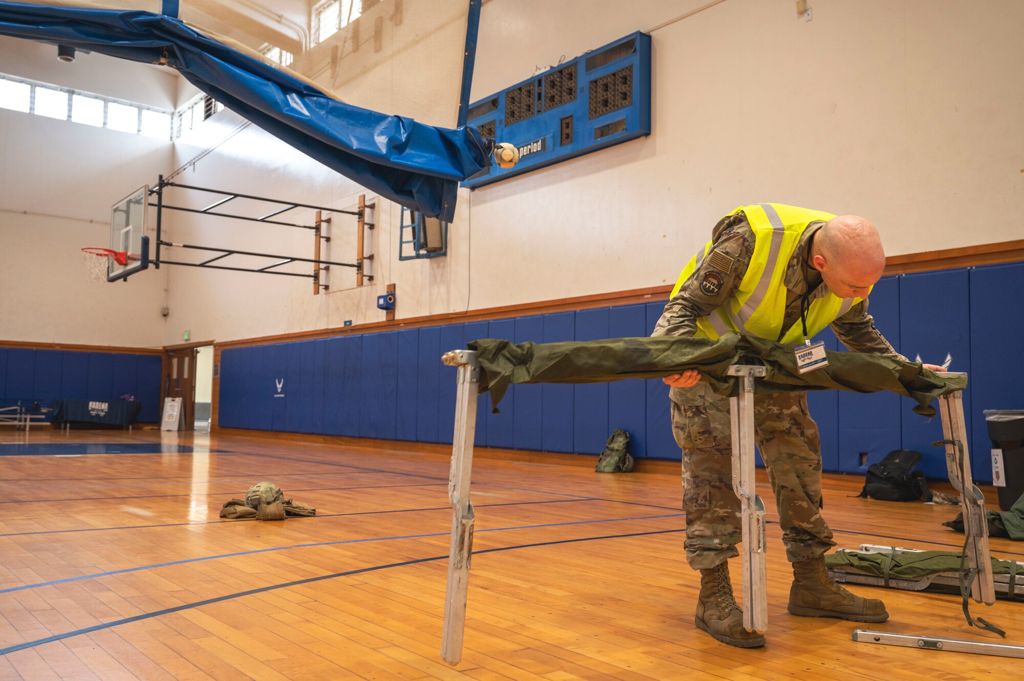 Air Force Member wears a reflective vest on a basketball court assembling a cot