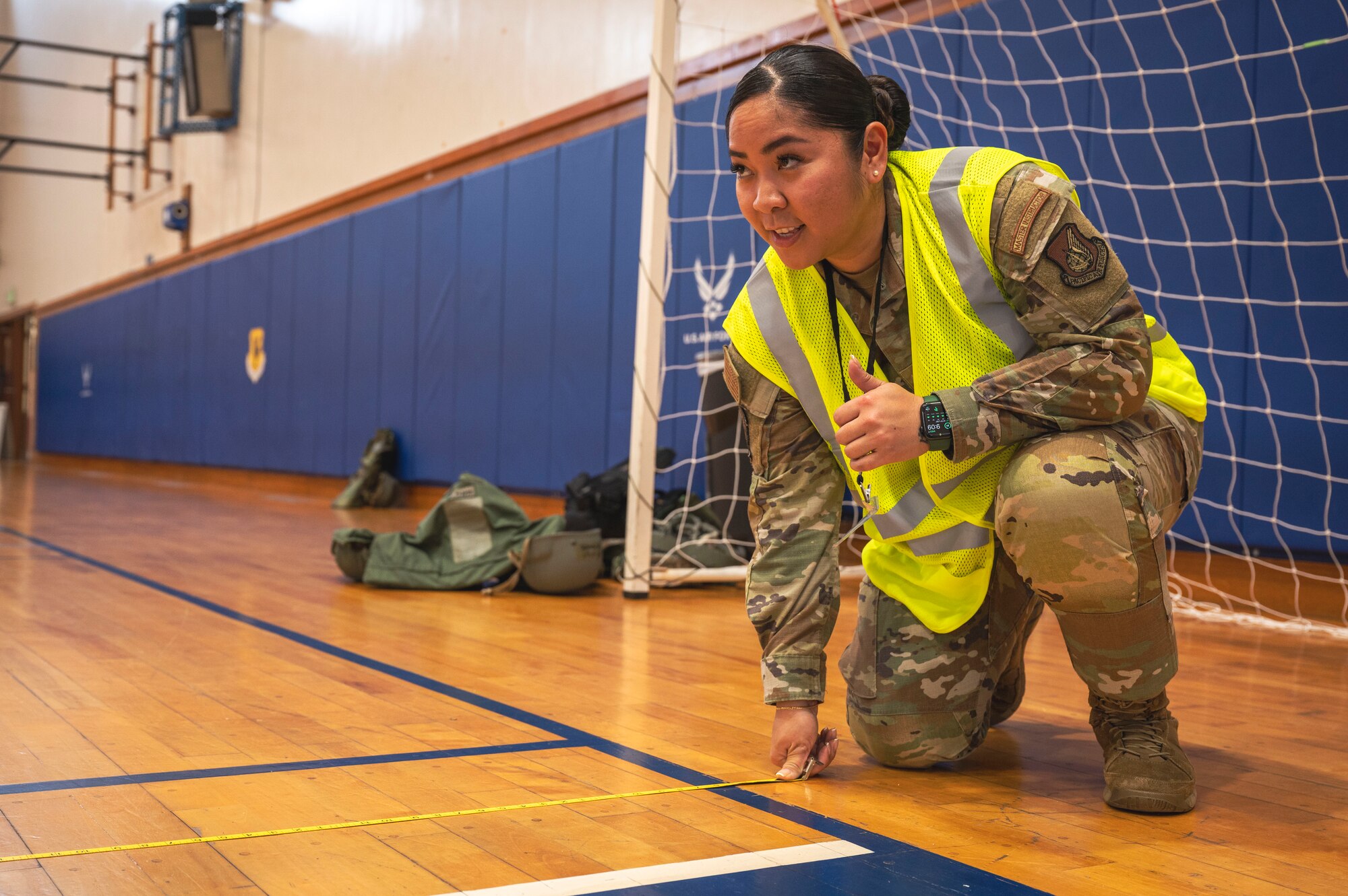 A Air Force Airman in a reflective vest kneals down on one knee on a indoor basketball court to use a tape measure