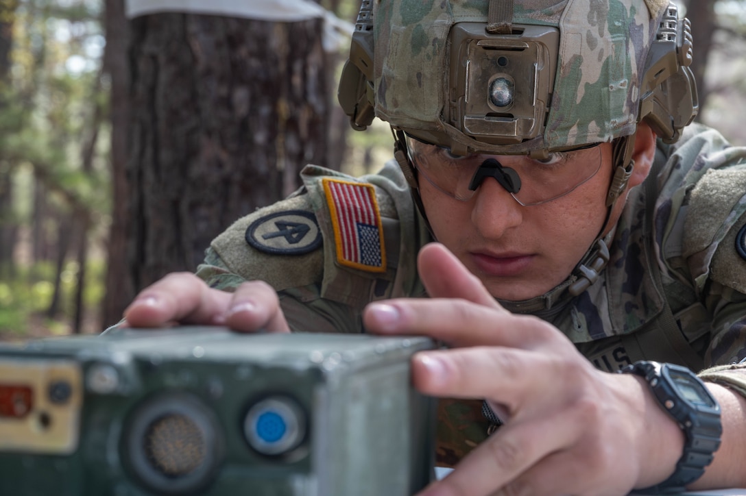 A close-up shows a soldier in tactical gear adjusting a radio near trees.