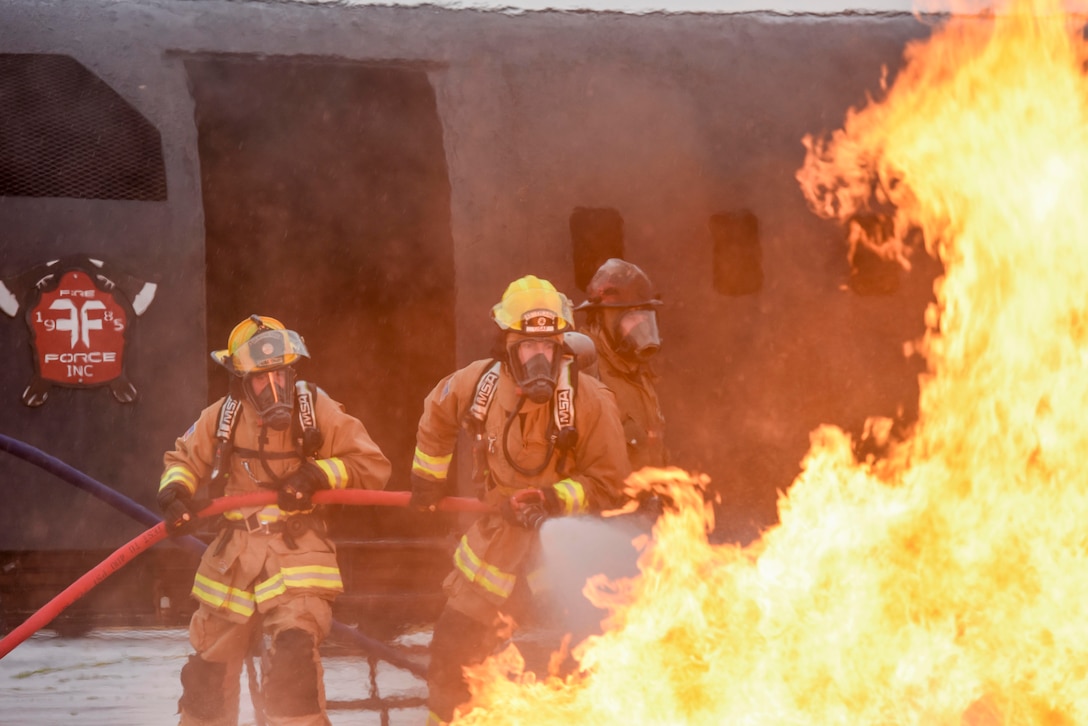 Three airmen in firefighting attire use a hose to extinguish flames during training.