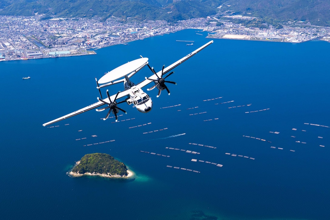 A Navy aircraft flies above blue waters with a small island, boats and a coastline in the distance.