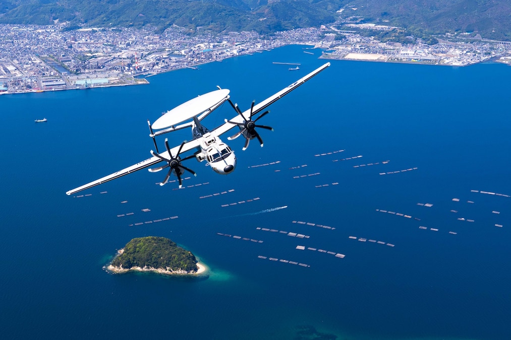 A Navy aircraft flies above blue waters with a small island, boats and a coastline in the distance.