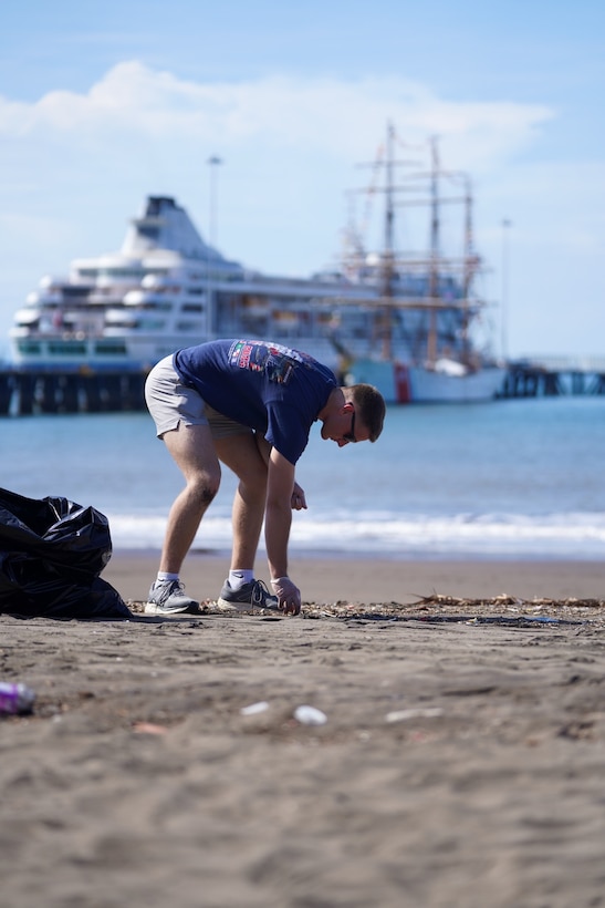 A crew member picks up trash on a local beach in Puntarenas, Costa Rica as the U.S. Coast Guard Cutter Eagle (WIX 327) sits moored in the background on April 27, 2025. Known as “America’s Tall Ship,” Eagle is a 295-foot, three-masted barque, which operates as a training vessel for future officers of the United States Coast Guard. (U.S. Coast Guard courtesy photo by U.S. Embassy Costa Rica.)