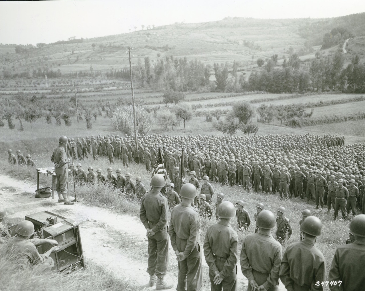 Hundreds of men in military uniforms and helmets stand in formation along a rolling hillside.