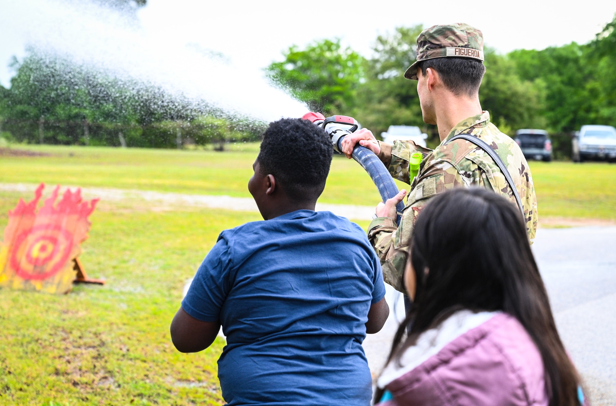 An Air Force firefighter demonstrates the function of a firehose to two students. The water sprays towards fake fire targets.