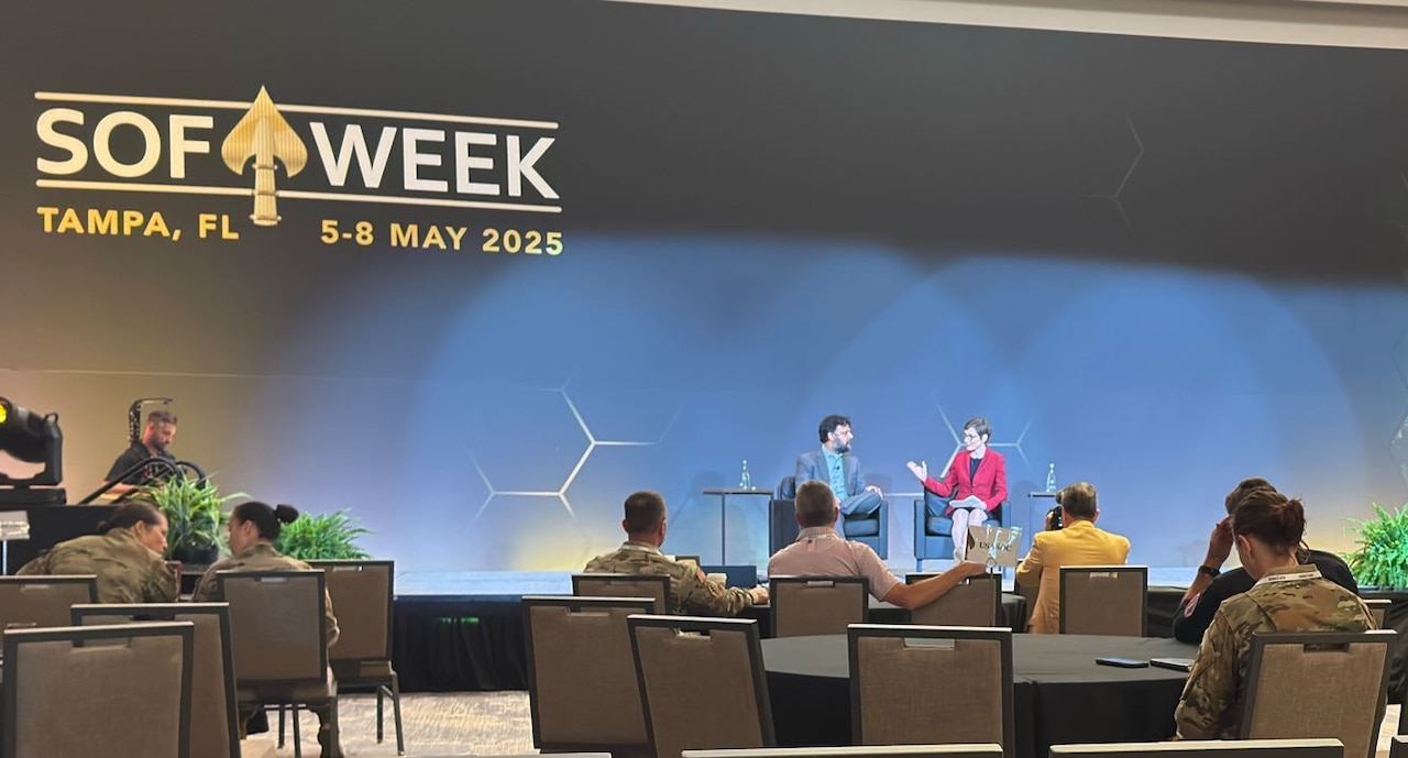 A man and a woman are sitting down having a discussion in a large room. A logo on the wall behind them and to the left reads “SOF WEEK, Tampa, FL, 5-8 May 2025.”