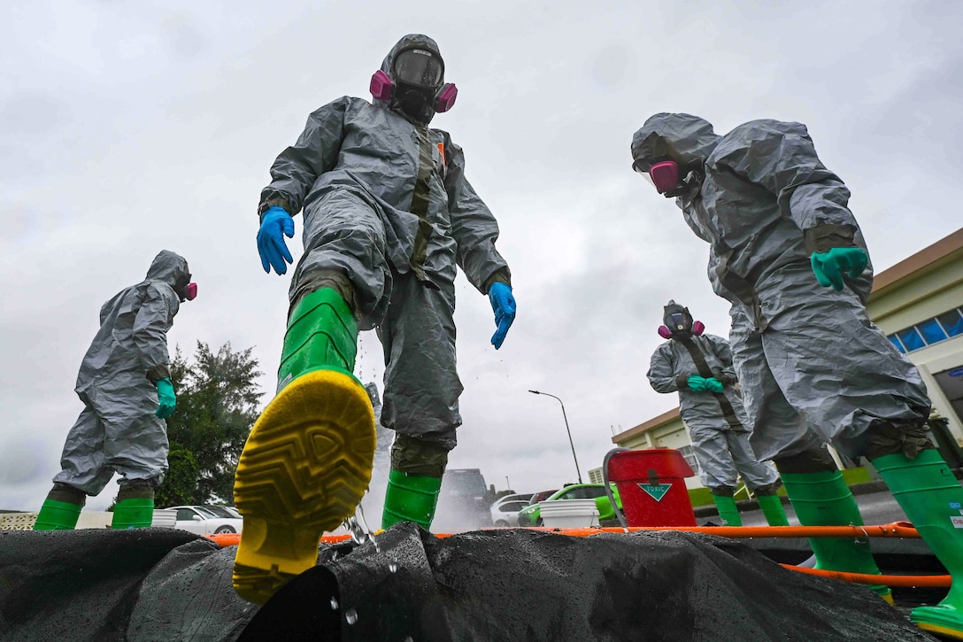 Airmen in decontamination suits stand outdoors as one steps forward on a black tarp on the ground.