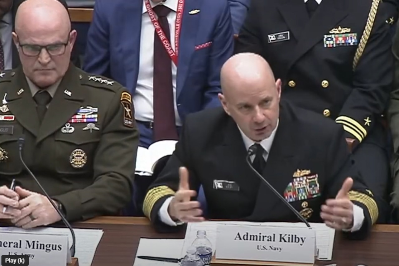 A man in a navy military uniform sits at a table with a microphone in front of his face. Several men in military uniforms and business attire sit beside and behind him.