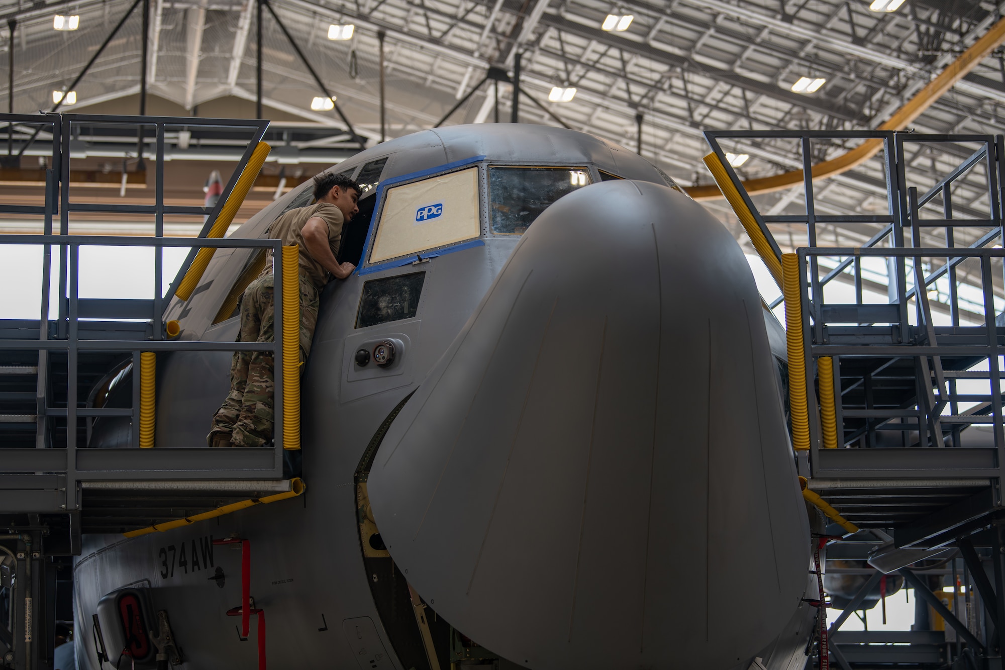 An Airman stands leans into a gray C-130J Super Hercules cockpit window from the outside.