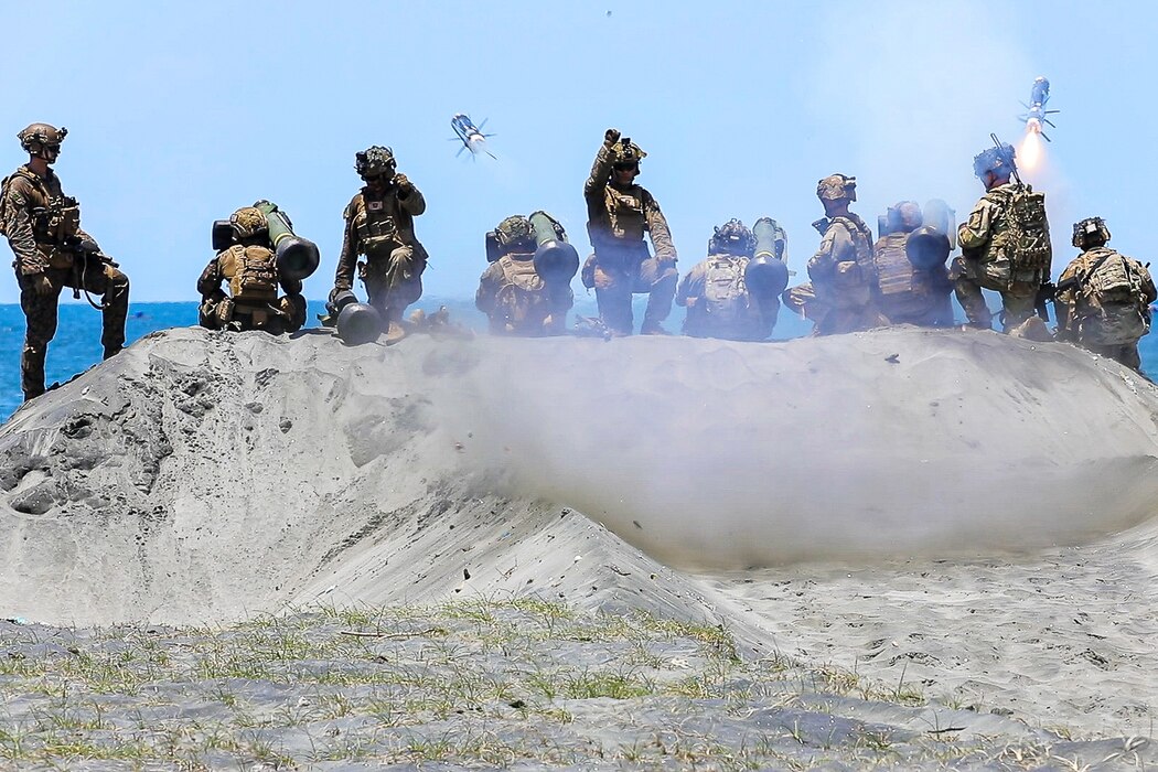 Ten soldiers and Marines on a sand dune fire two missiles toward a body of water during daytime.
