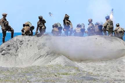 Ten soldiers and Marines on a sand dune fire two missiles toward a body of water during daytime.