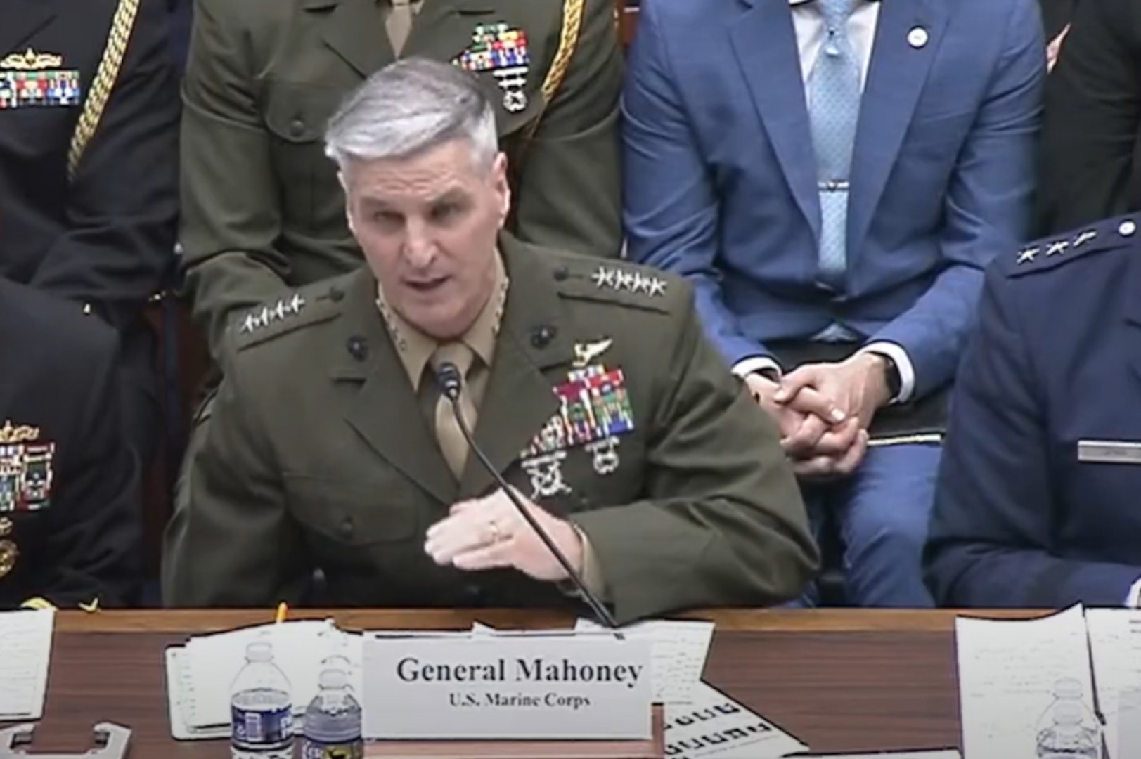A man in a green military uniform sits at a table while speaking into a microphone. Several men in military uniforms and business attire sit beside and behind him.