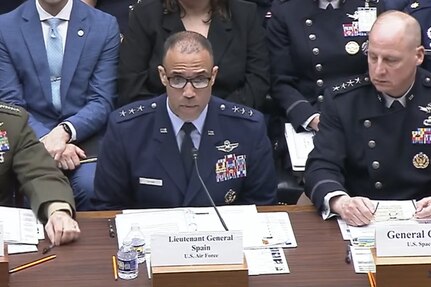 A man in a blue military uniform sits at a table while speaking into a microphone. Several men in military uniforms and business attire sit beside and behind him.