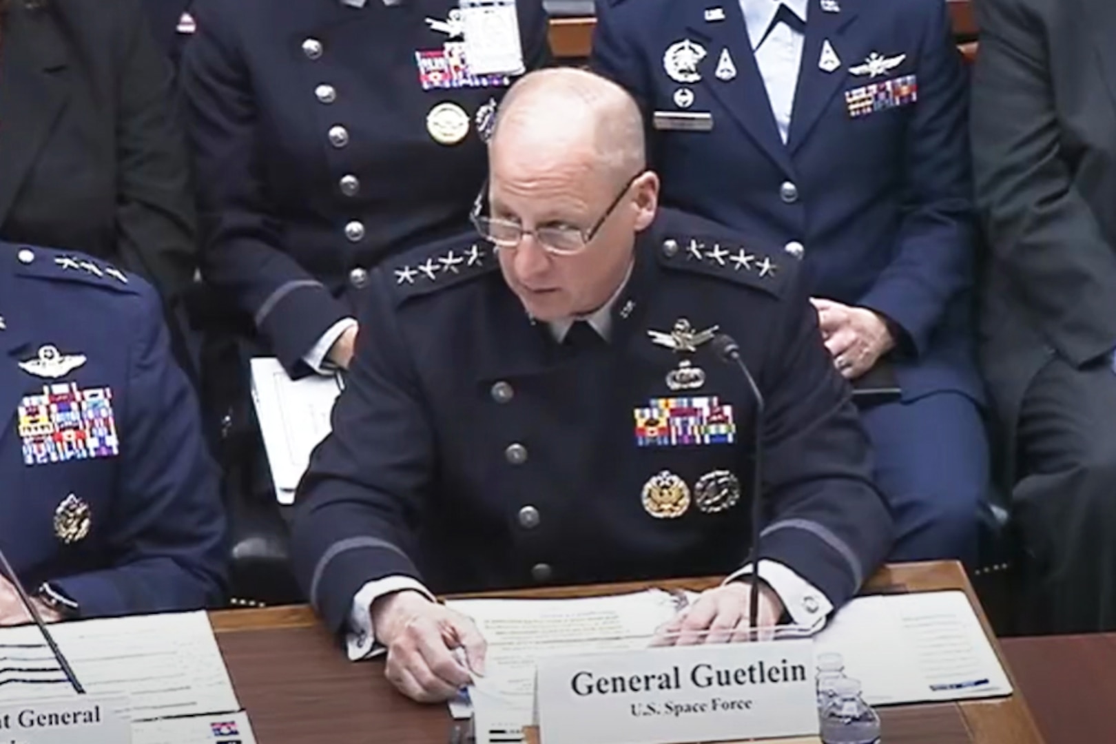 A man in a blue military uniform sits at a table while speaking into a microphone. Several men in military uniforms and business attire sit beside and behind him.
