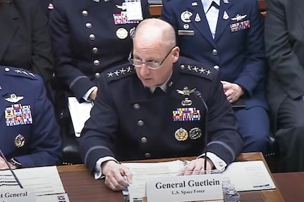 A man in a blue military uniform sits at a table while speaking into a microphone. Several men in military uniforms and business attire sit beside and behind him.