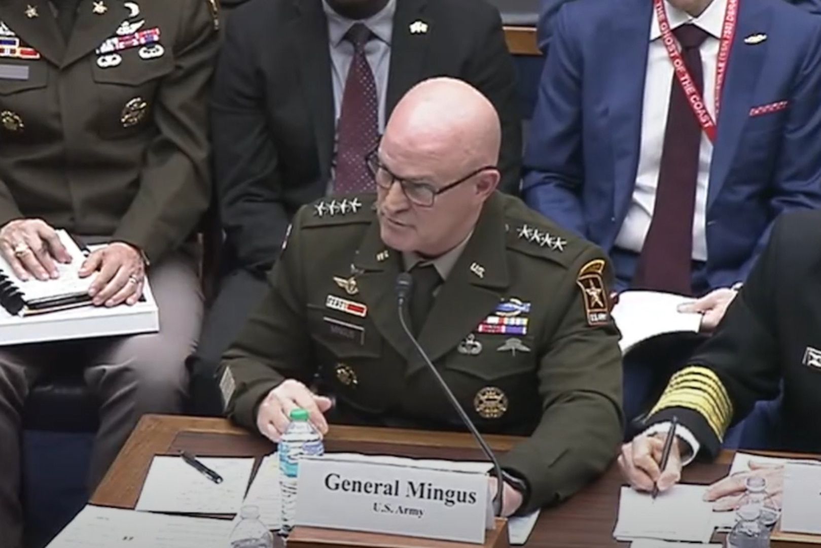 A man in a green military uniform sits at a table speaking into a microphone. Several men in military uniforms and business attire sit beside and behind him.