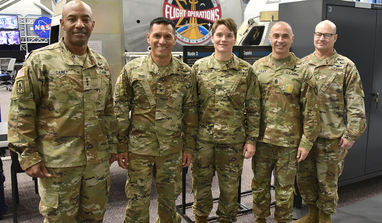 Four men and a woman wearing camouflage military uniforms pose for a photo inside a large room; there is equipment in the background and a command logo that reads, "Flight Operations."