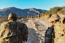 Soldiers conducting a patrol in Greece on a dirt road with a mountain range in front of them.
