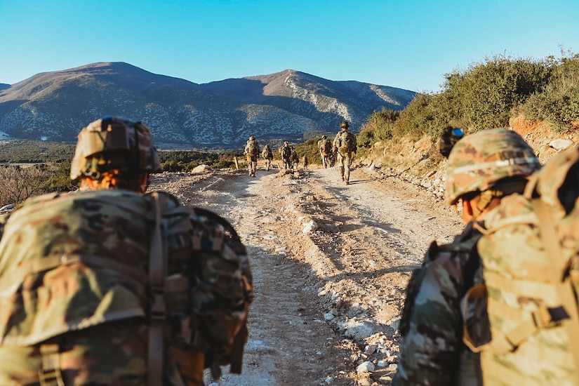 Soldiers conducting a patrol in Greece on a dirt road with a mountain range in front of them.