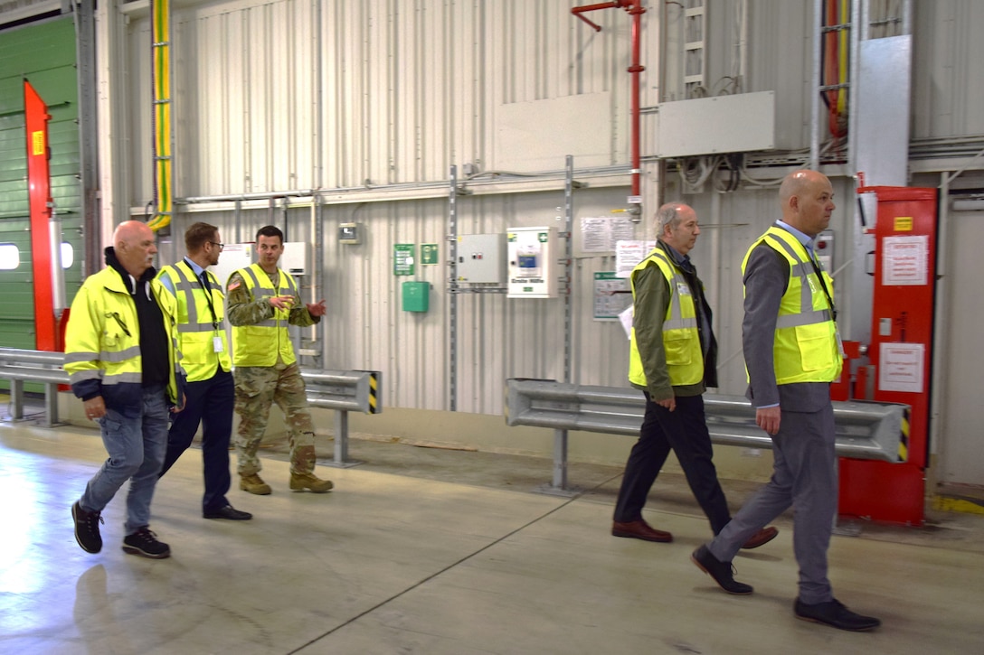 A group of people wearing reflective safety vests walk through a warehouse talking to one another.