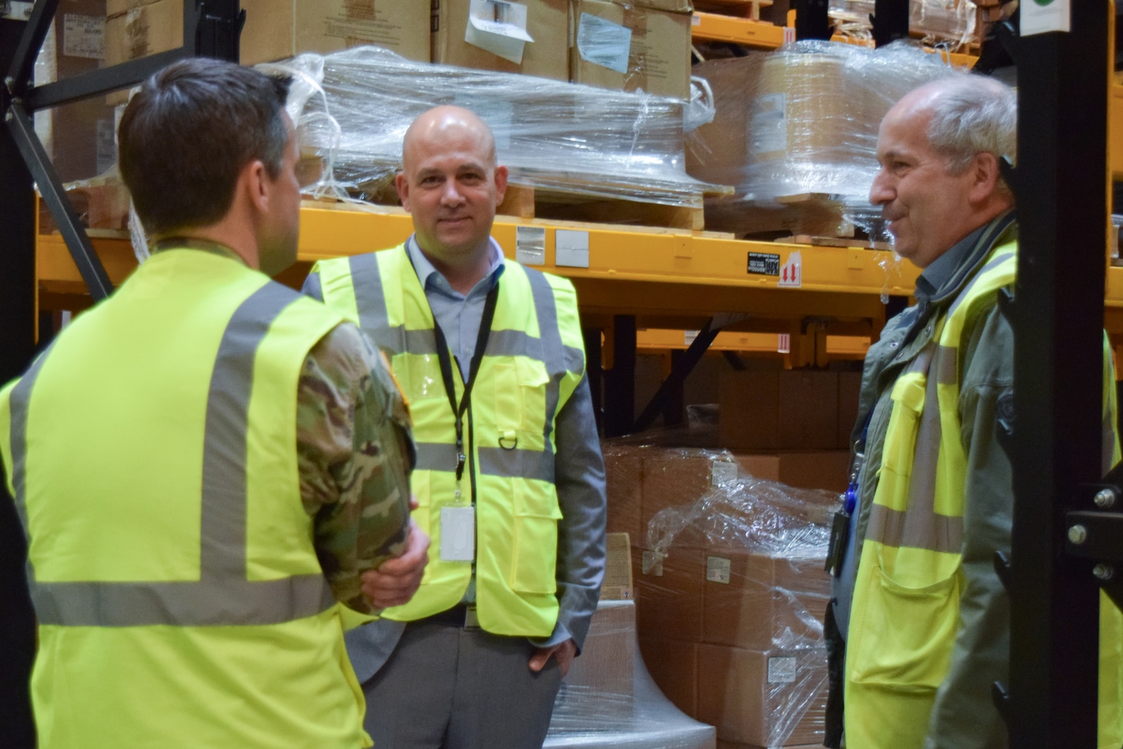 Three men wearing reflective safety vests stand in front of shelving in a warehouse talking to one another.