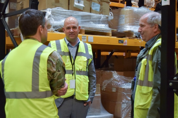 Three men wearing reflective safety vests stand in front of shelving in a warehouse talking to one another.