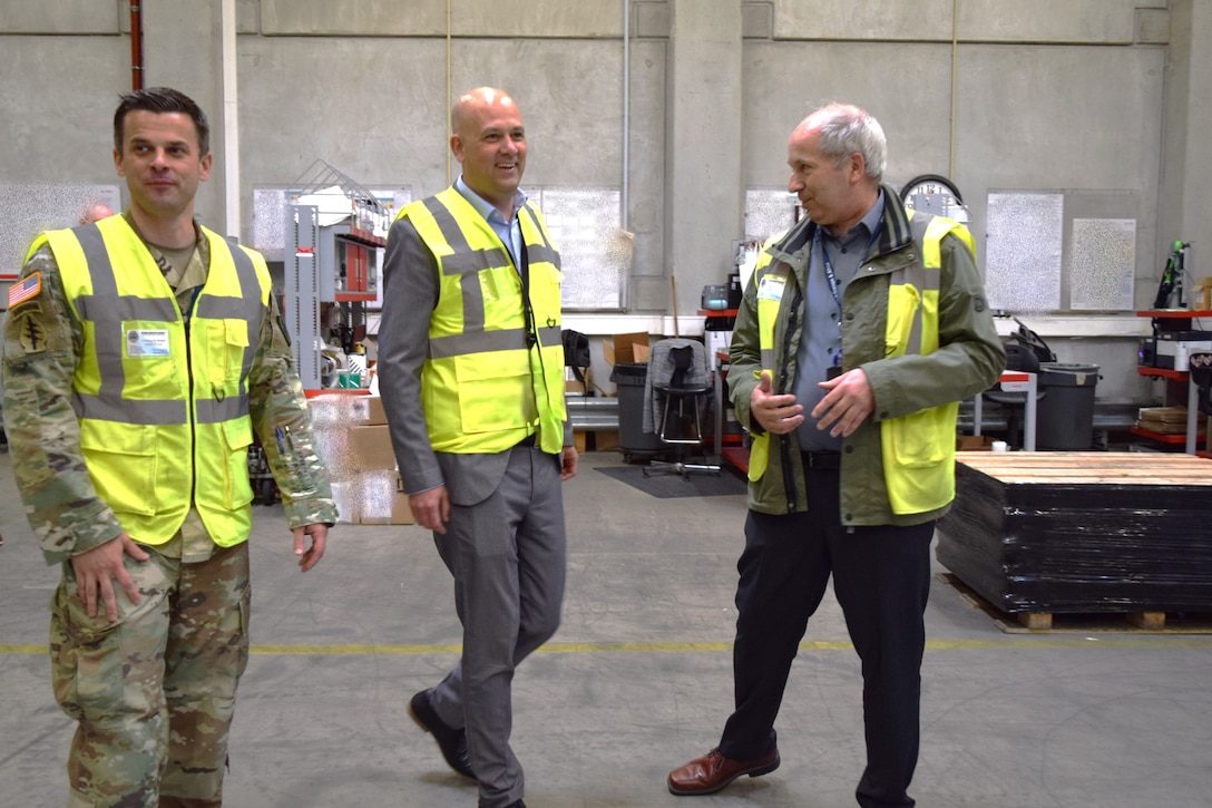 Three men wearing reflective safety vests walk through a warehouse talking to one another.