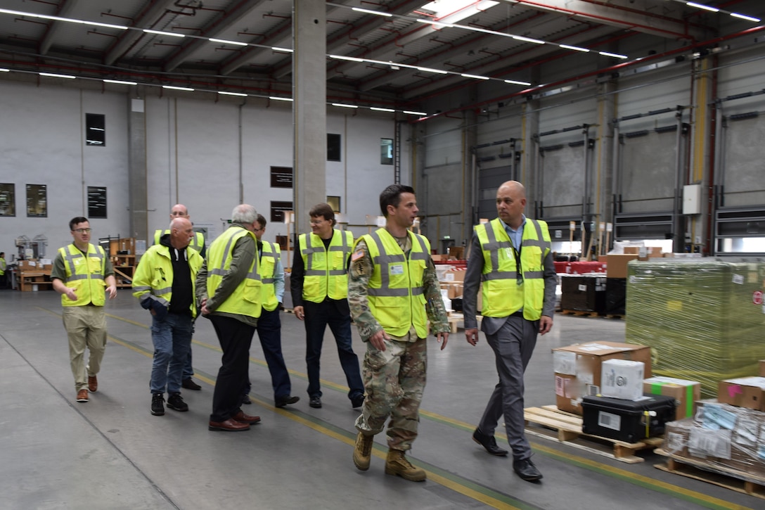 A group of people wearing reflective safety vests walk through a warehouse talking to one another.