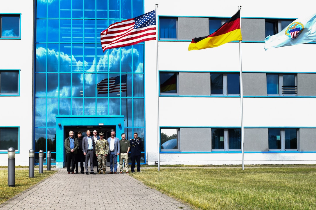 A group of people stand in front of a large building on a sunny day with the American, German and Defense Logistics Agency flags flying in front of the building.